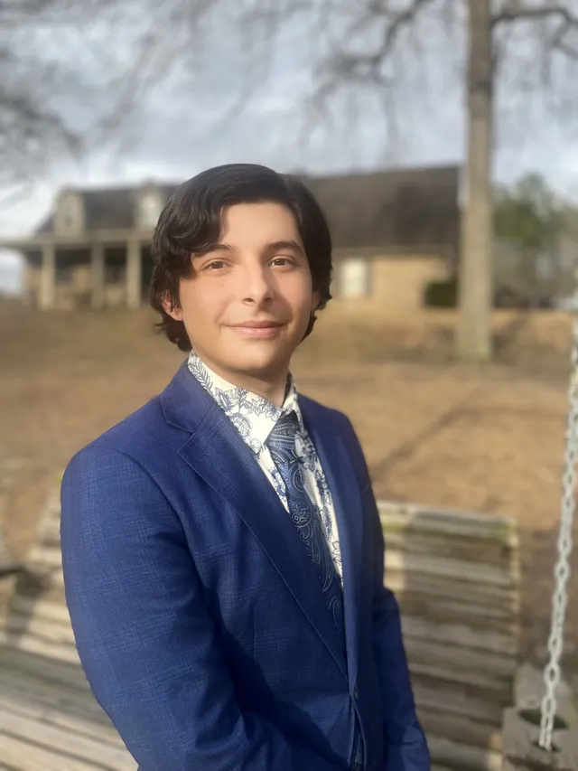 Jorge Naranjo Jr in a navy suit standing outdoors beside a wooden porch swing