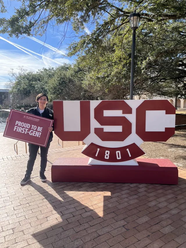Jorge Naranjo Jr holding a “Proud to be First-Gen!” sign beside the USC 1801 sign on campus